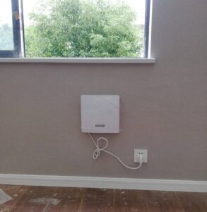 Alternative text: White Telmann wireless alarm receiver mounted on beige wall with power plug, close to window with greenery outside.